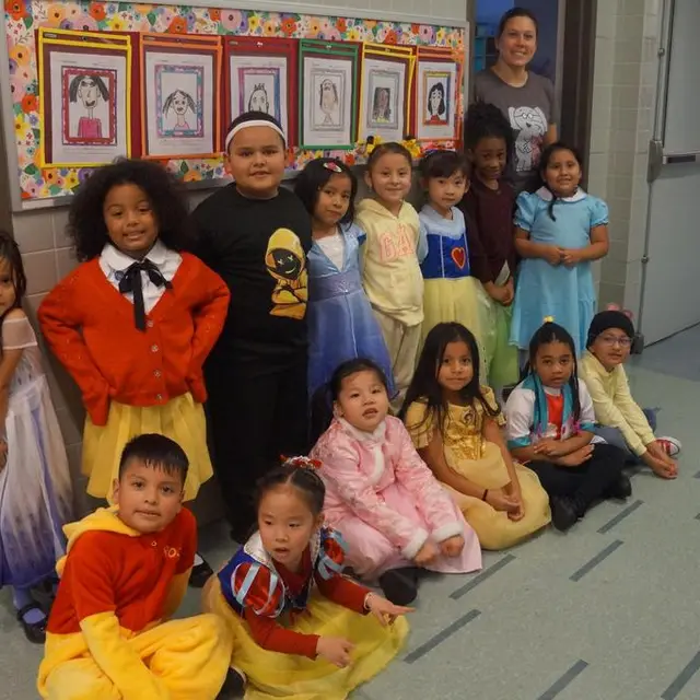 A class in costume seated on the rug with their teachers behind them