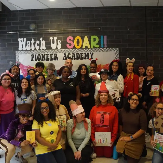 Staff in costume holding books in front of the “Watch Us SOAR!! Pioneer Academy” wall