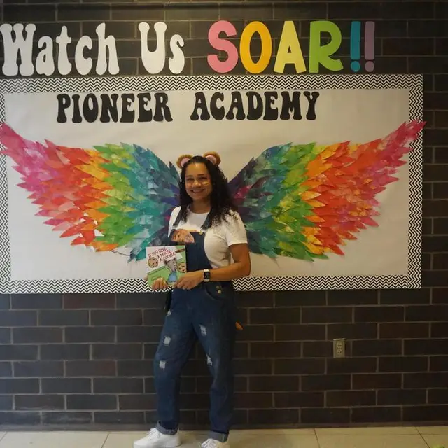 A staff member in overalls holding a book in front of the rainbow wings mural