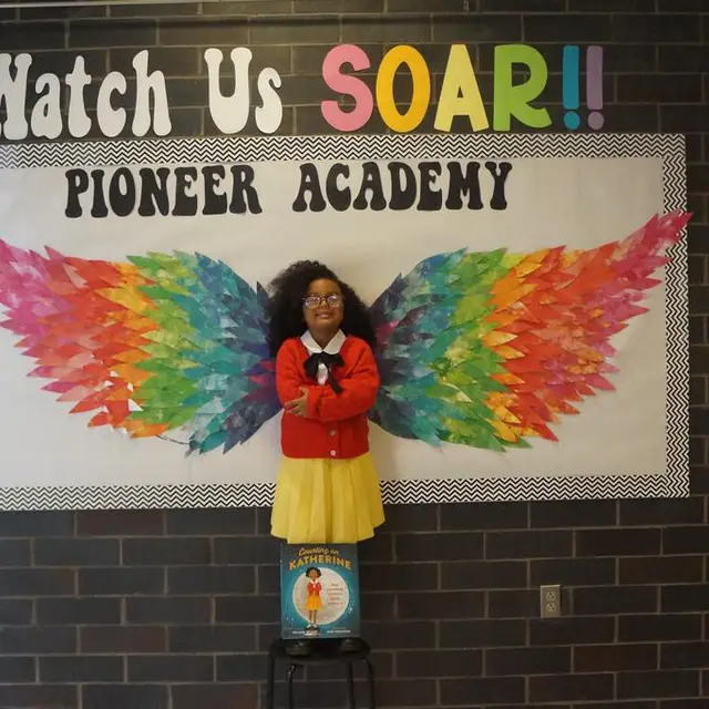 A scholar in a red jacket and yellow skirt posing with a book in front of the rainbow wings mural