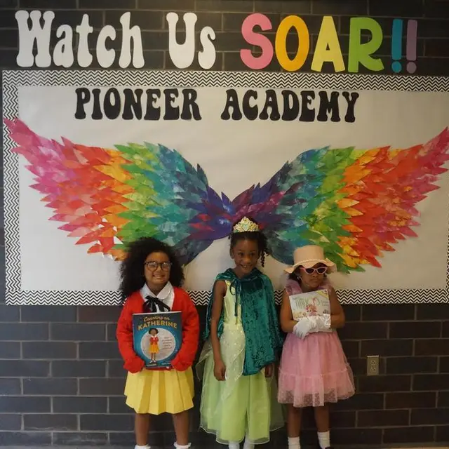 Three scholars in costume holding books in front of the rainbow wings mural