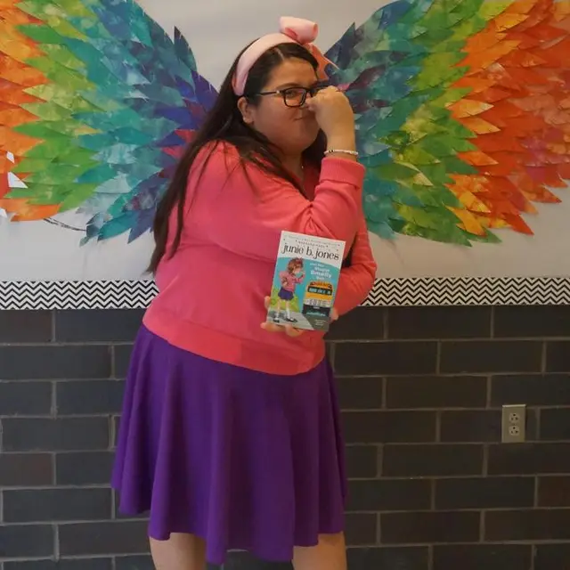 A staff member in a pink sweater and purple skirt holding the book “Junie B. Jones”