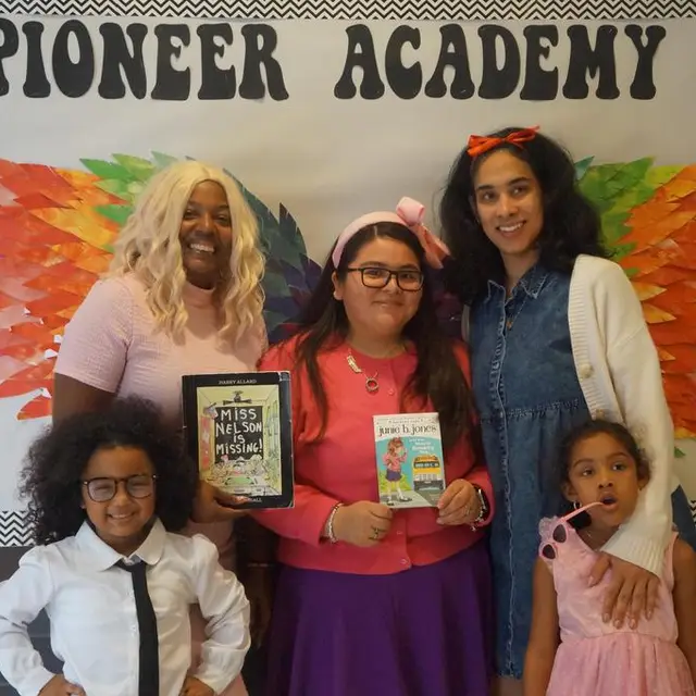 Staff and scholars in costume holding books in front of the Pioneer Academy wall
