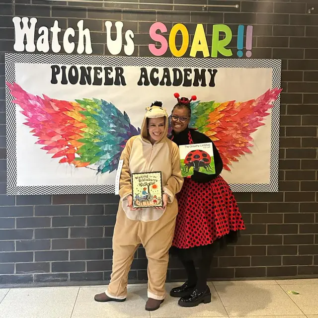 Two staff members dressed as storybook animals holding books in front of the rainbow wings mural